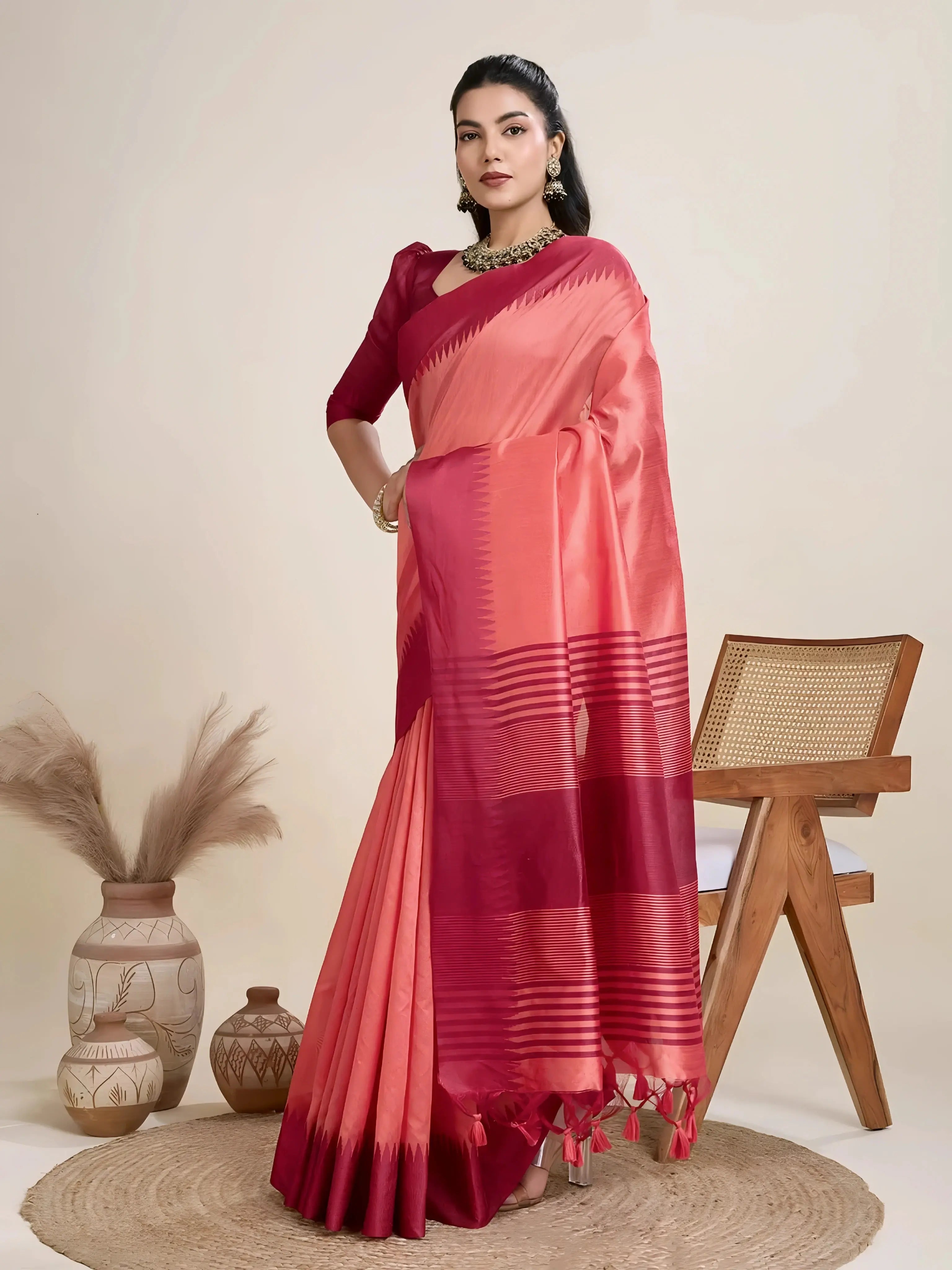 A woman models a striking two-toned Raw Silk Saree. The main body is Coral Pink, and the pallu/border contrast dramatically in Deep Maroon/Wine Red. The saree features traditional temple weaving and horizontal stripe patterns on the pallu. She wears a long-sleeve maroon blouse and accessorizes with a chunky dark gemstone necklace, posing indoors.